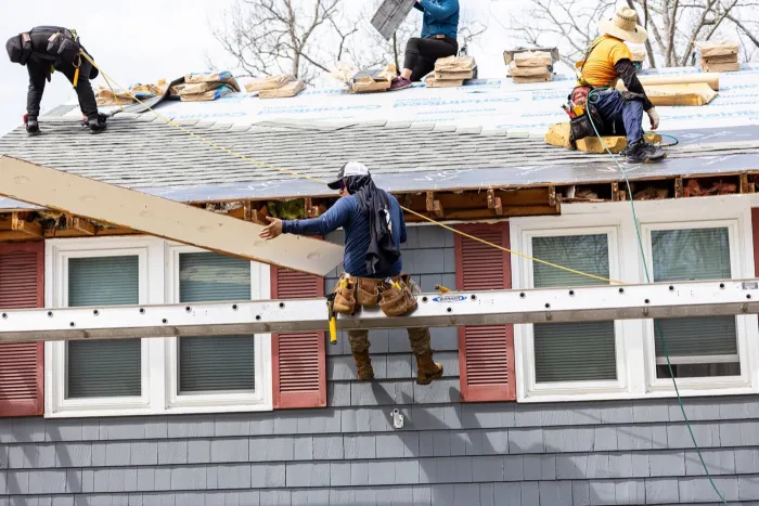 Senco crew passing a long board along the open eave of a gray-sided home during a roof replacement
