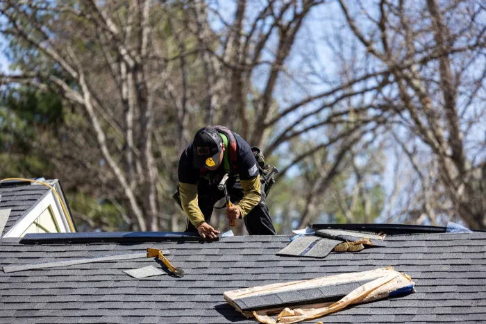 Senco roofer working along the ridge of an asphalt shingle roof, with shingle pieces and a bundle wrapper beside him