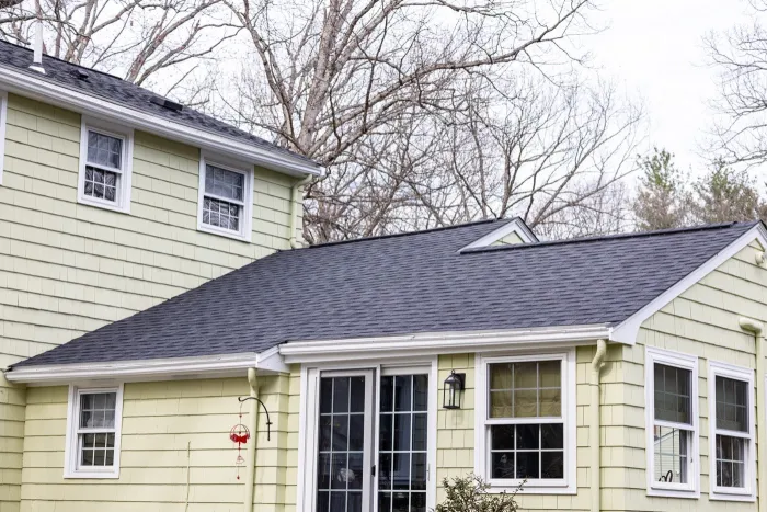 Finished dark asphalt shingle roof on a pale yellow home, with bare trees behind it