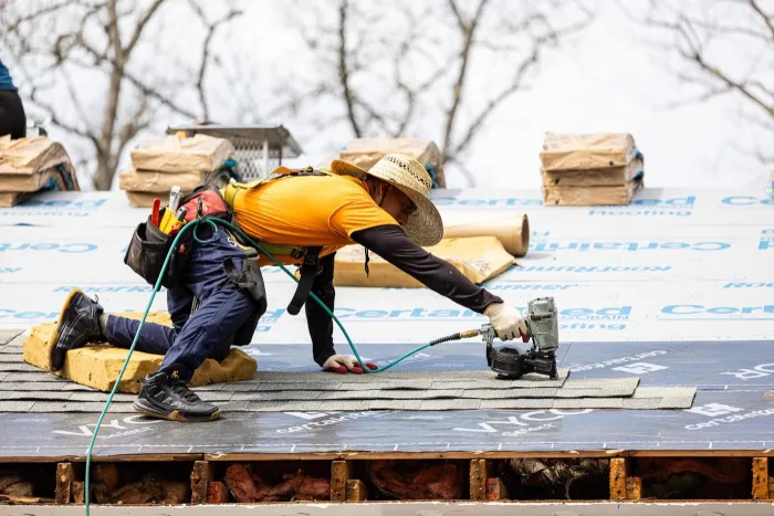 Senco roofer nailing shingles over new CertainTeed underlayment, with the open eave showing rafters and attic insulation below